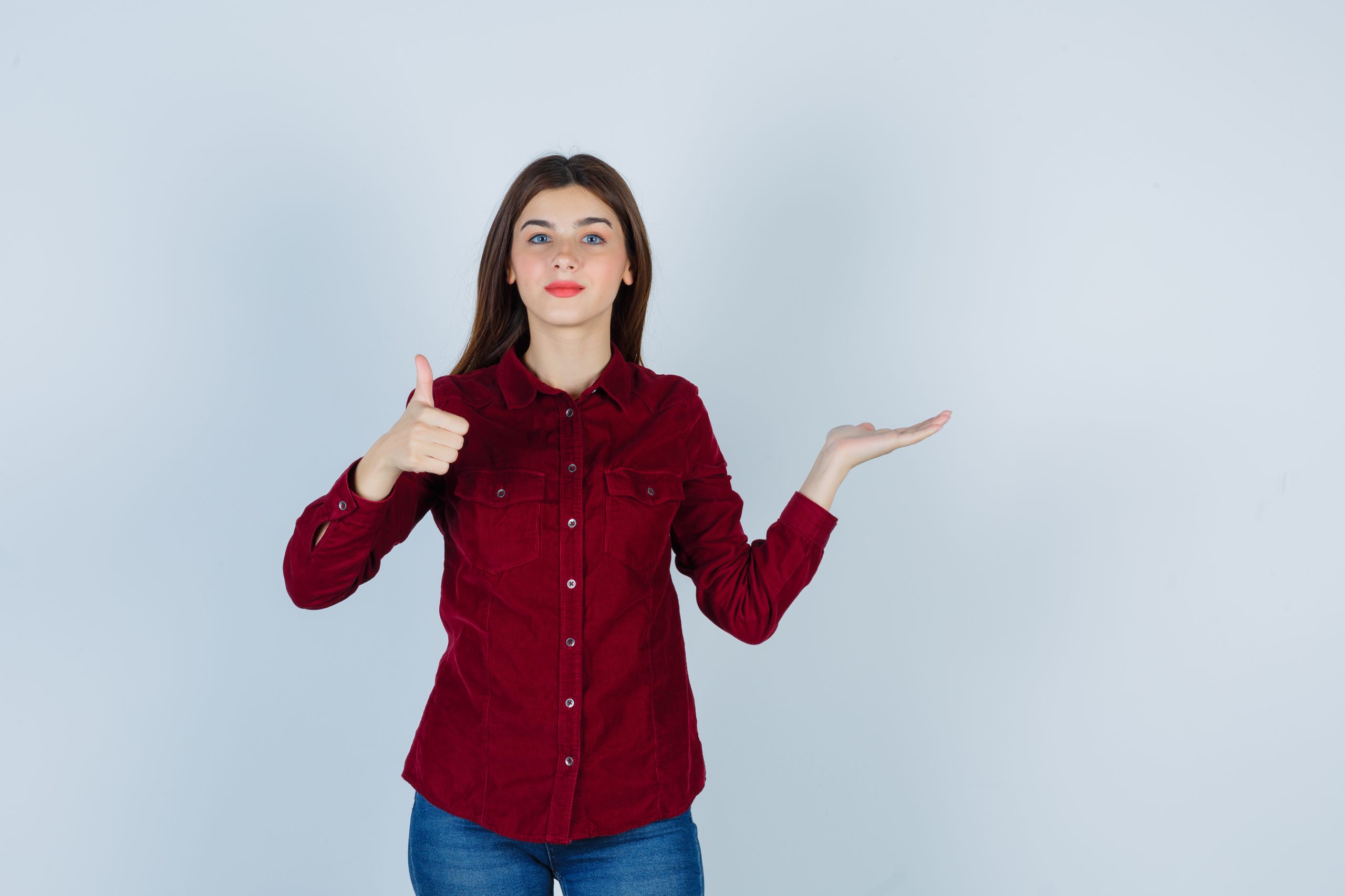 portrait of teenage girl showing thumb up, spreading palm aside in burgundy shirt and looking confident front view